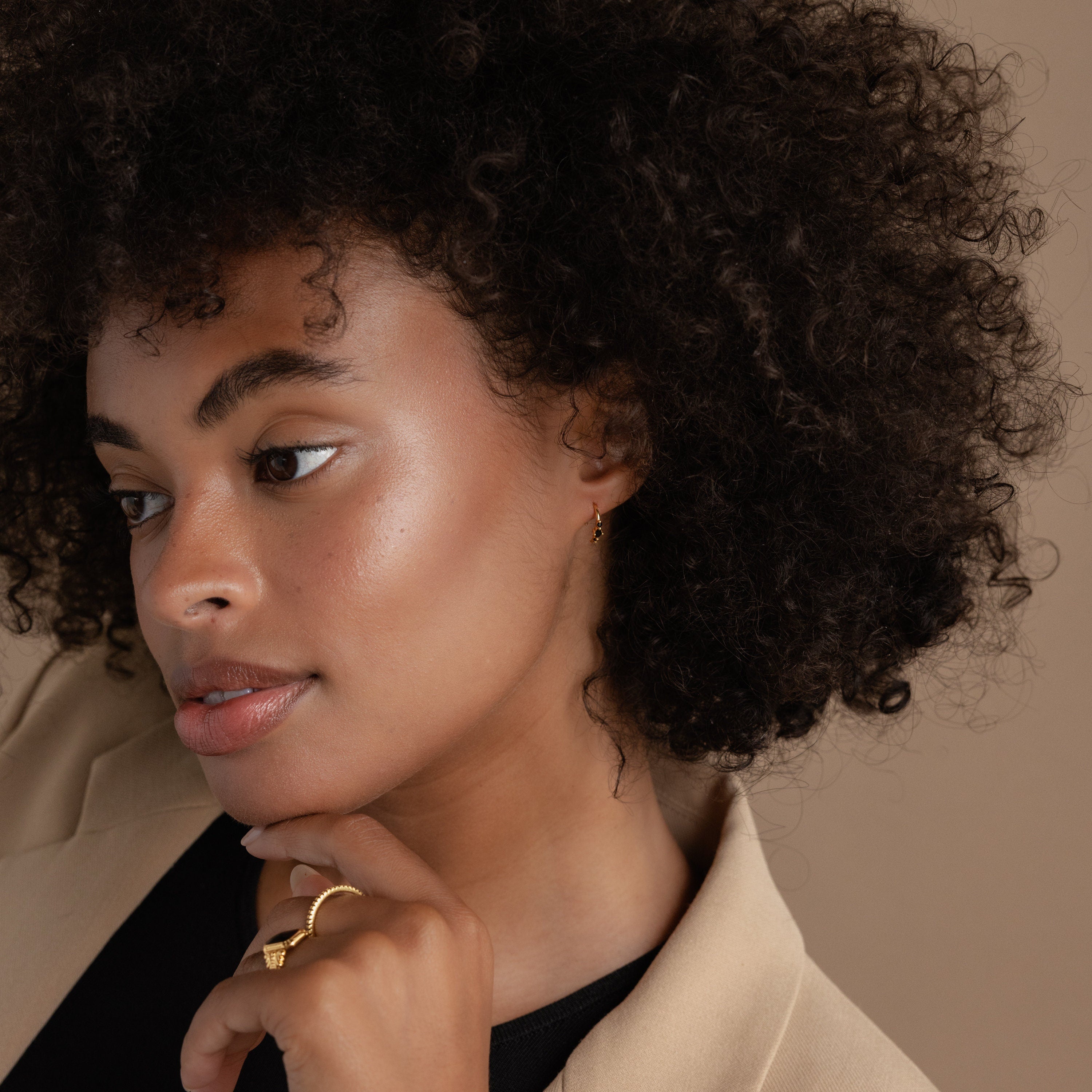 Woman with curly hair in a beige blazer, looking to the side with hand near her chin, wears gold pieces from the latest Jewelry Collection and striking Amaya Black Marquise Threaders.