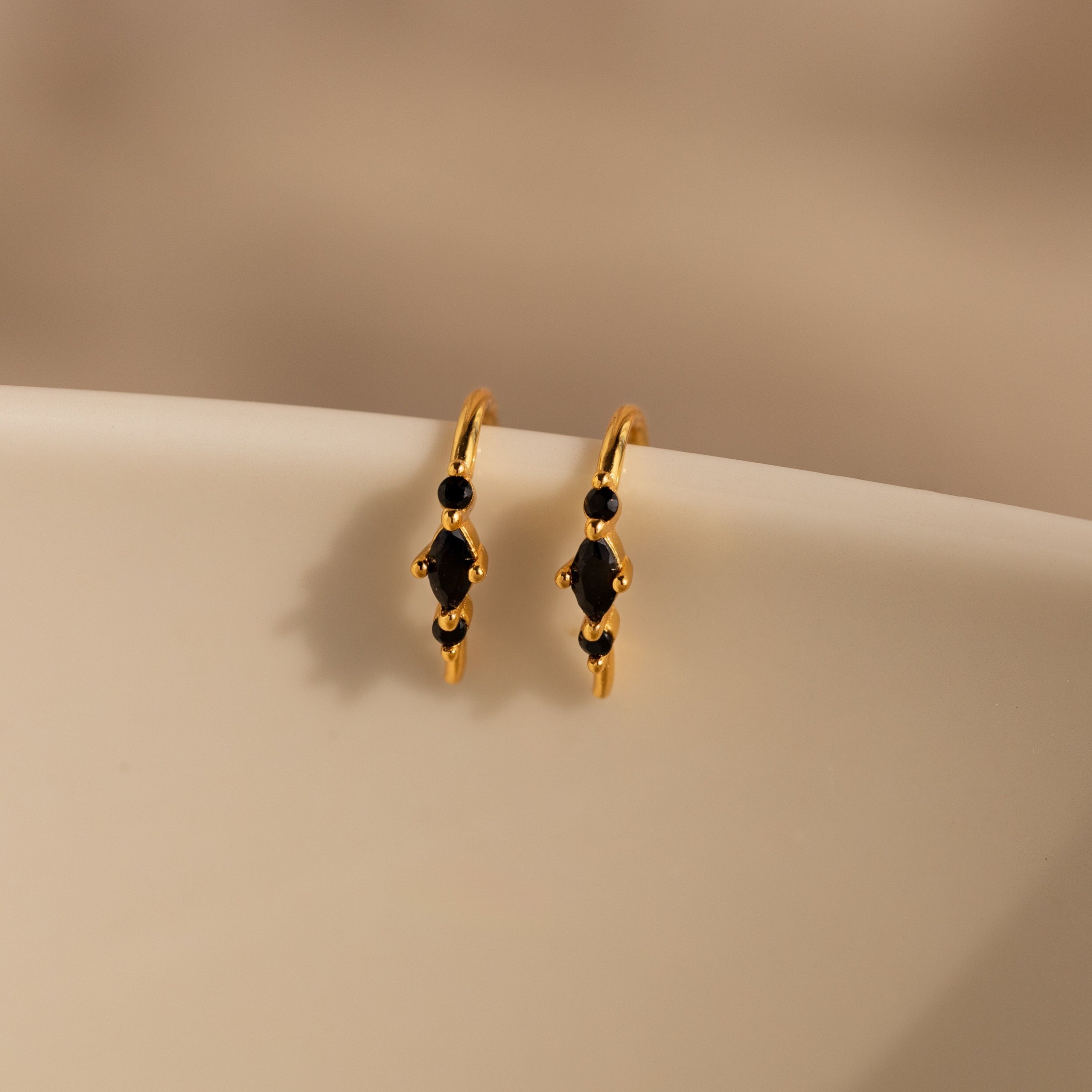 The Amaya Black Marquise Threaders, featuring gold hoops with black gemstone accents, rest on a smooth cream-colored surface—a stunning addition to any modern earrings collection.