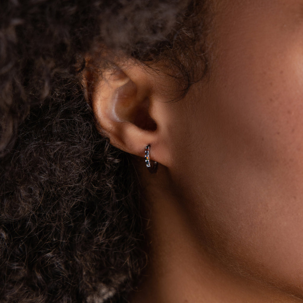 Close-up of a person’s ear wearing Black Sand Opal Huggies—small, silver hoops with embedded opal gemstones, perfect as everyday earrings.