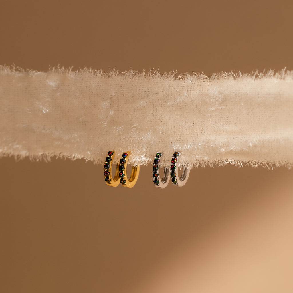 Two dainty hoop earrings—one gold, one silver—decorated with colorful gems rest on a soft surface. The Black Sand Opal Huggies are perfect as everyday earrings, adding a vibrant touch to any look.