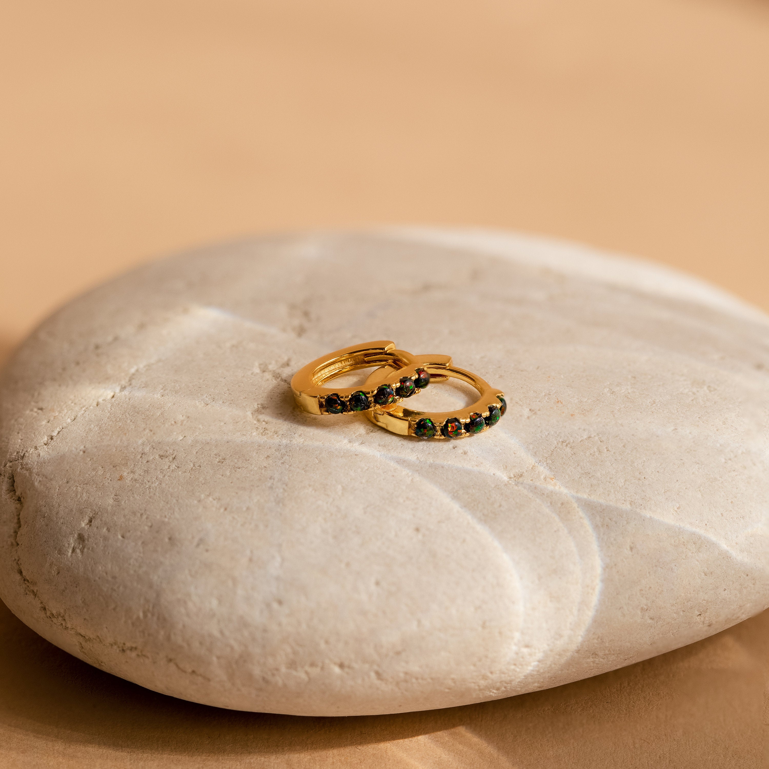 The Black Sand Opal Huggies feature gold hoops with dark green stones, displayed on a smooth, light-colored surface—ideal dainty earrings for everyday wear.