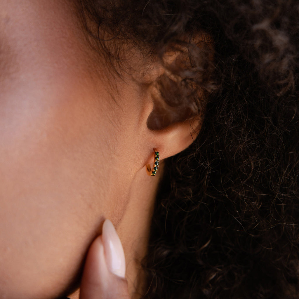 Close-up of a woman's ear wearing Black Sand Opal Huggies, dainty hoop earrings with green stones; her hand rests near her face.