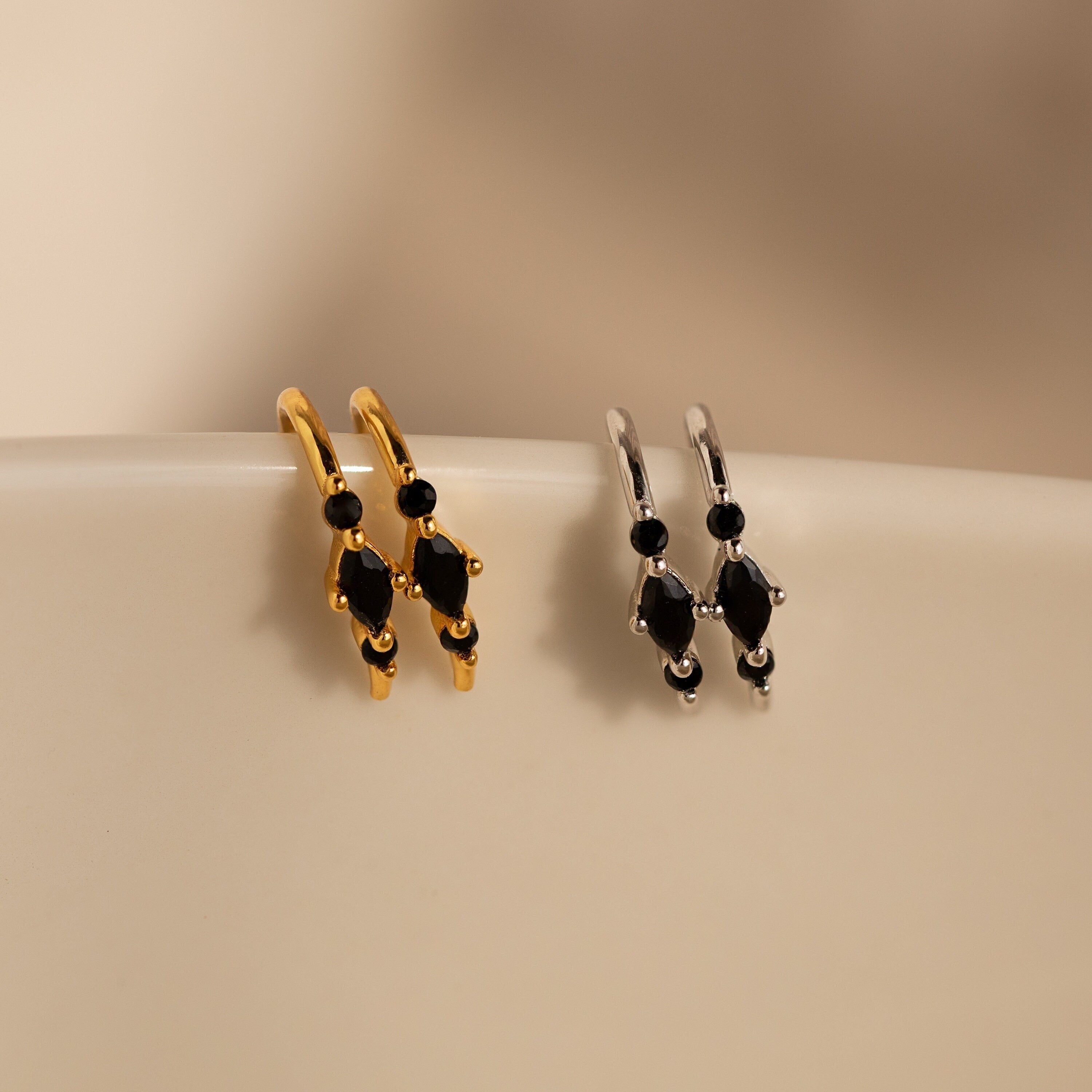 Amaya Black Marquise Threaders—gold and silver modern earrings with black stones, displayed on a smooth cream surface—make an elegant addition to any jewelry collection.