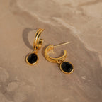 Black Gemstone Drop Hoops are shown on a beige marble surface, offering a chic update to everyday earrings with their gold hoops and striking black gemstone charms.
