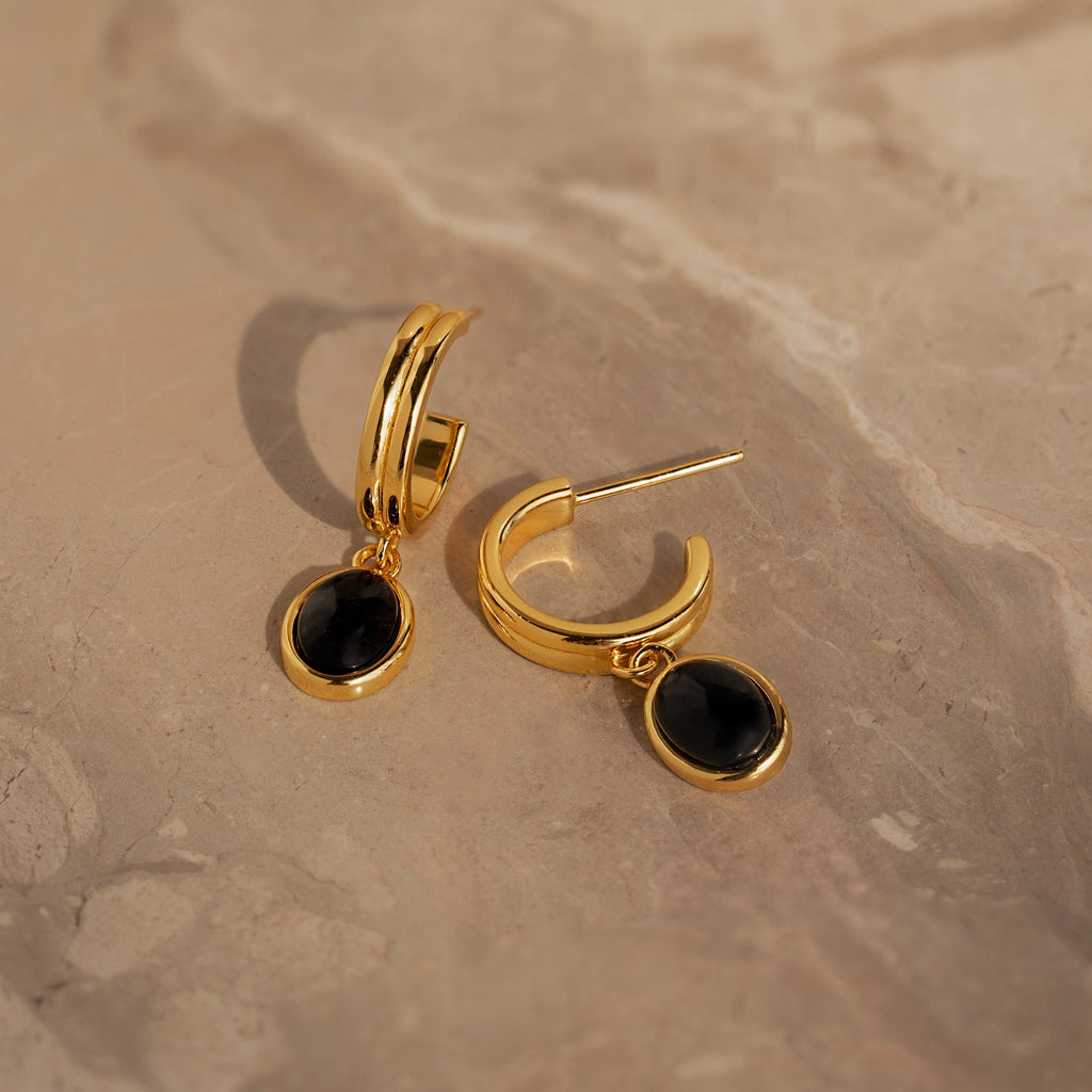 The Onyx Diamond Earrings Set features gold hoops with oval black stones, adding a touch of elegance when displayed on a beige marble surface.