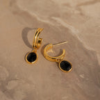 The Onyx Diamond Earrings Set features gold hoops with oval black stones, adding a touch of elegance when displayed on a beige marble surface.