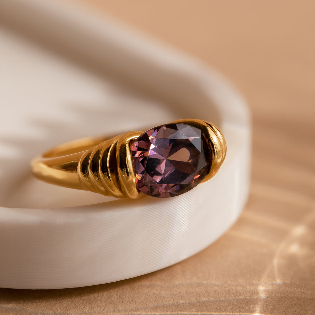 The Dark Amethyst Signet Ring, featuring a large purple gemstone in an art deco gold setting, rests on a white ceramic dish with a warm background.