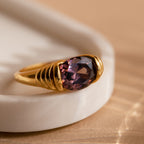 The Dark Amethyst Signet Ring, featuring a large purple gemstone in an art deco gold setting, rests on a white ceramic dish with a warm background.