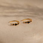 The Amaya Black Marquise Threaders feature gold hoops with black stone accents, elegantly displayed on a beige textured surface—an exquisite addition to any jewelry collection.