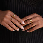 Two hands with manicured nails wear the Black Emerald Ring and gold bands, set against a black ribbed fabric background.