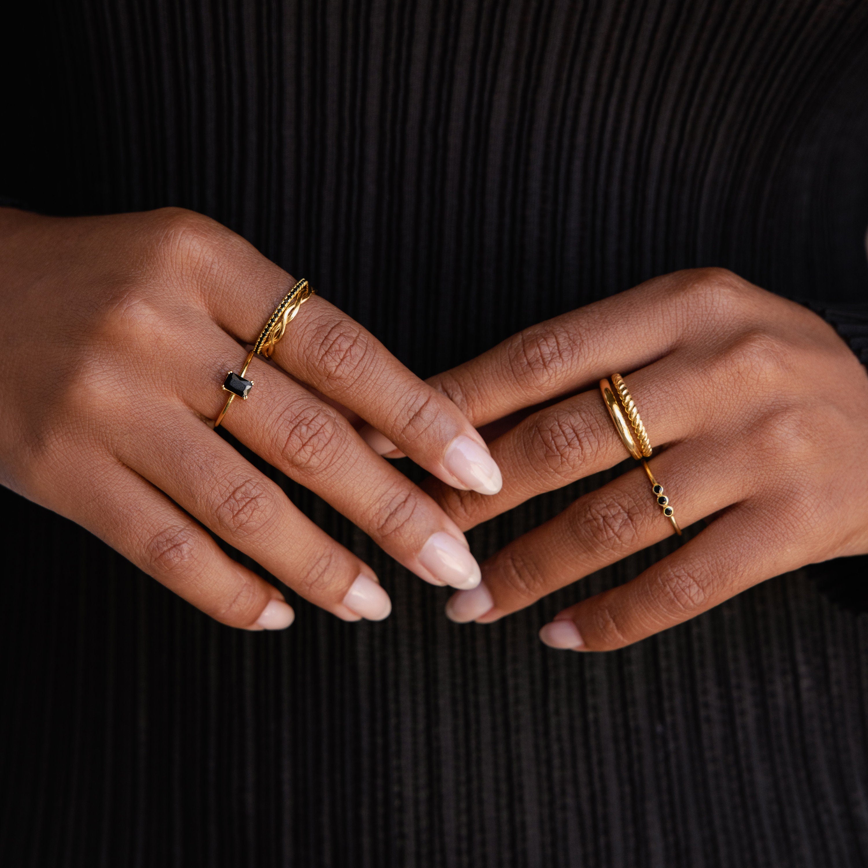 Two hands with manicured nails wear the Black Emerald Ring and gold bands, set against a black ribbed fabric background.