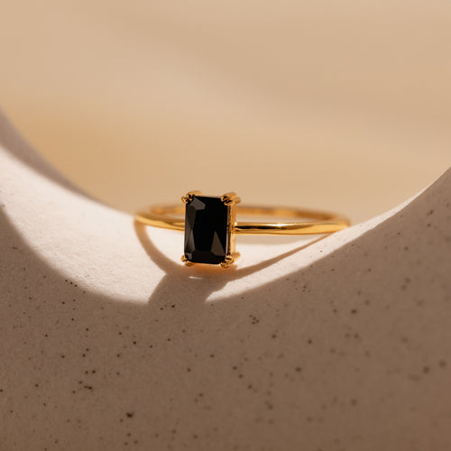 The Black Emerald Ring—a gold Art Deco piece with a rectangular black gemstone—evokes vintage-inspired elegance, showcased on a smooth, light-colored surface.