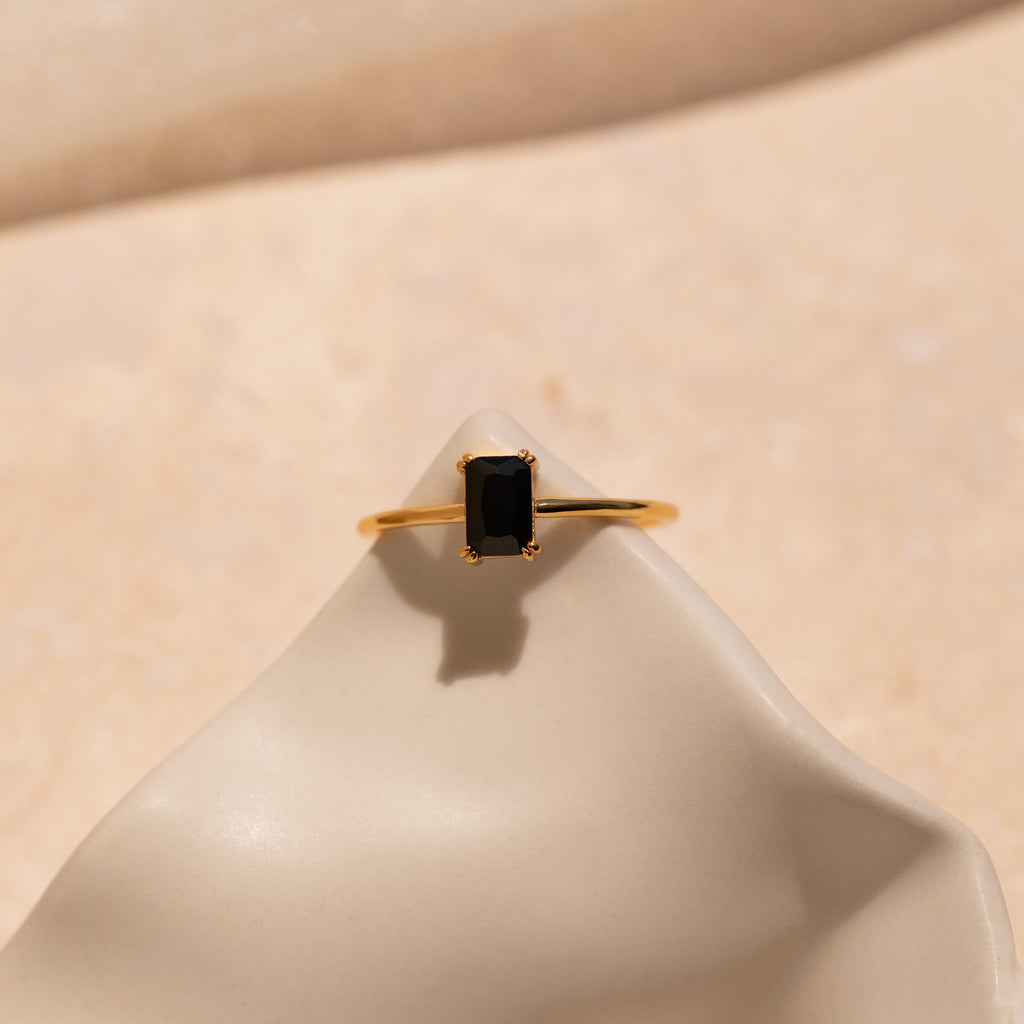 The Black Emerald Ring is a gold band with a rectangular black stone inspired by Art Deco style, shown elegantly on a smooth, cream-colored surface.
