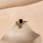 The Black Emerald Ring is a gold band with a rectangular black stone inspired by Art Deco style, shown elegantly on a smooth, cream-colored surface.