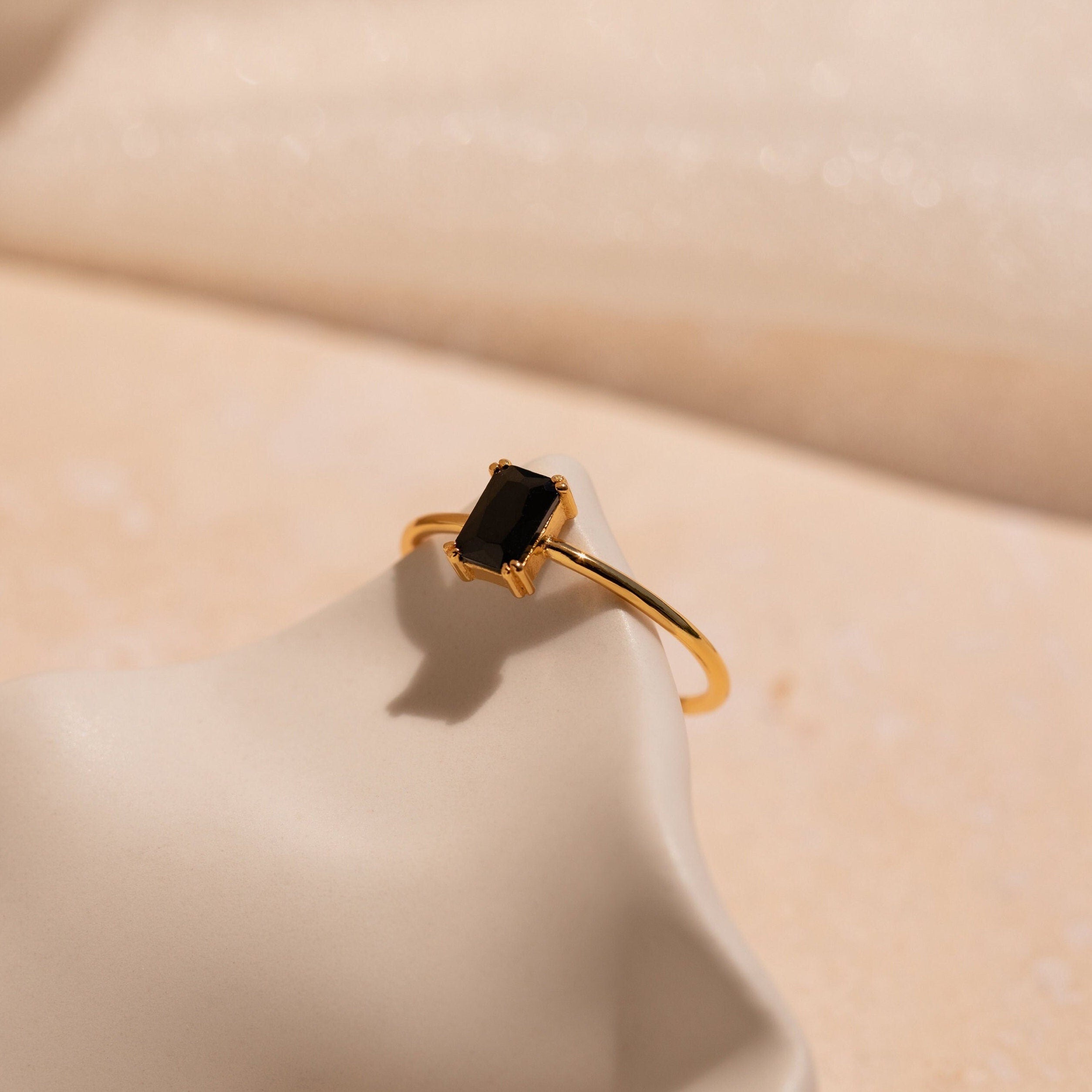 The Black Emerald Ring, featuring a rectangular black onyx stone in a vintage Art Deco design, rests elegantly on a smooth cream-colored surface under soft lighting.