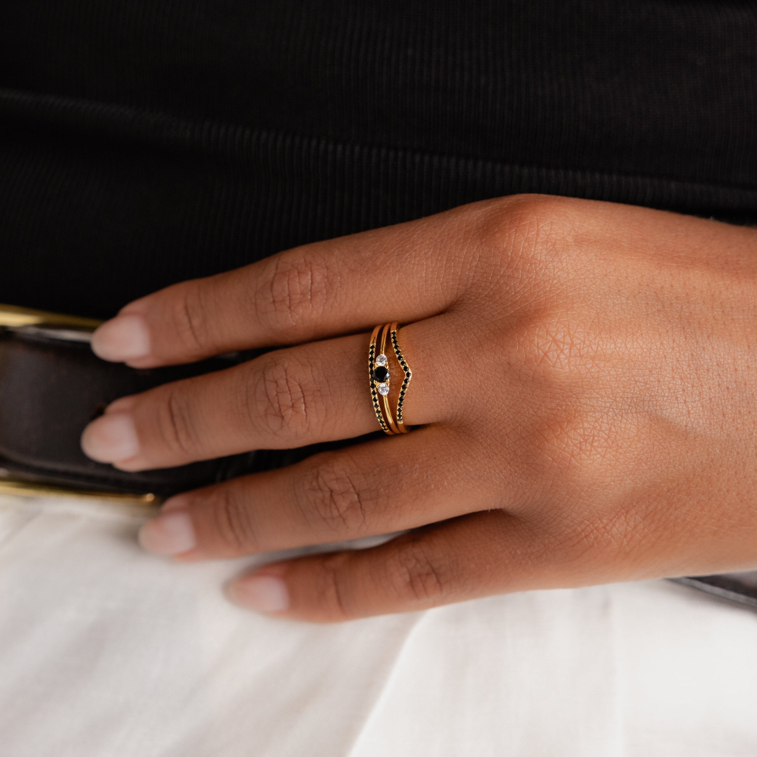 A hand with manicured nails wears the elegant Black Curve Ring Set, featuring a gold band with black and white stones, resting gracefully on a white skirt.