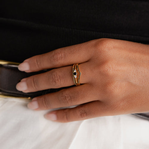 A hand with manicured nails wears the elegant Black Curve Ring Set, featuring a gold band with black and white stones, resting gracefully on a white skirt.