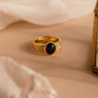 The Celestial Black Signet Ring features a midnight blue sandstone oval stone and gold finish, displayed on soft beige fabric—a stunning statement jewelry piece.