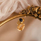 The Celestial Black Signet Ring, featuring a gold wavy band and a dark oval stone, is displayed on an ornate gold mirror atop soft fabric—a striking statement jewelry piece.