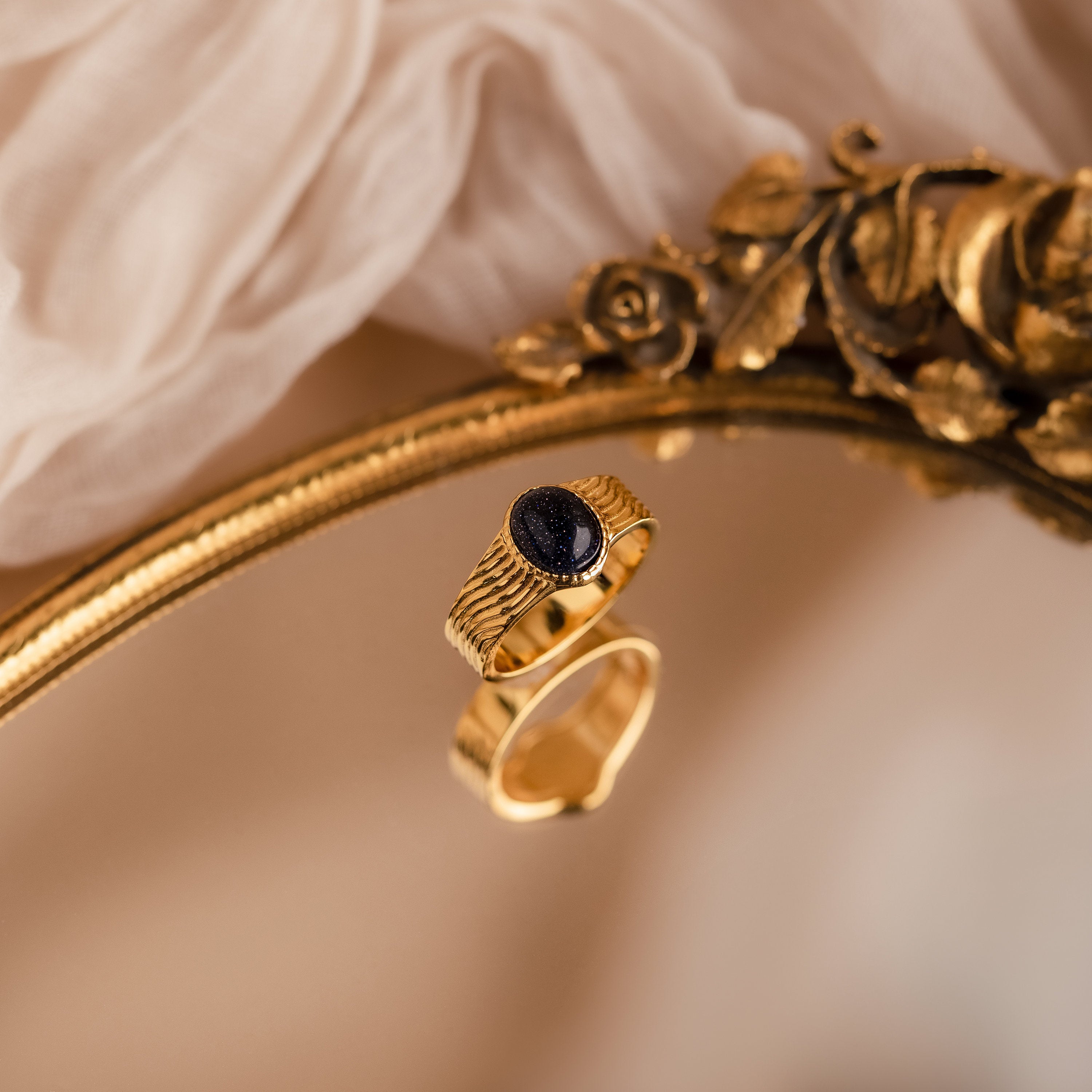 The Celestial Black Signet Ring, featuring a gold wavy band and a dark oval stone, is displayed on an ornate gold mirror atop soft fabric—a striking statement jewelry piece.