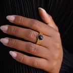 A hand with manicured nails wears a Celestial Black Signet Ring, showcasing its striking black oval stone as statement jewelry against dark fabric.