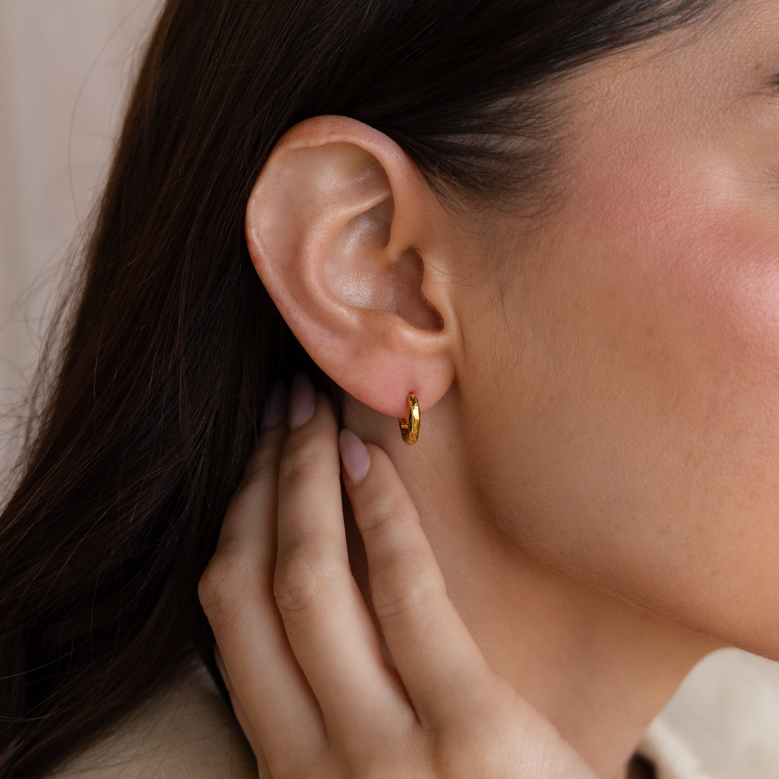 Close-up of a woman's ear adorned with the Mini Hammered Hoops Set; her hand gently touches her neck, highlighting the elegance of these gold earrings.