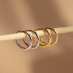 The Mini Bold Hoops Set—featuring one silver and one gold pair—hangs elegantly on a light wooden stick against a beige background, highlighting the timeless beauty of classic hoop earrings.