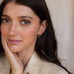 A woman with long brown hair gently touches her face while wearing the Mini Hammered Hoops Set and a beige top.