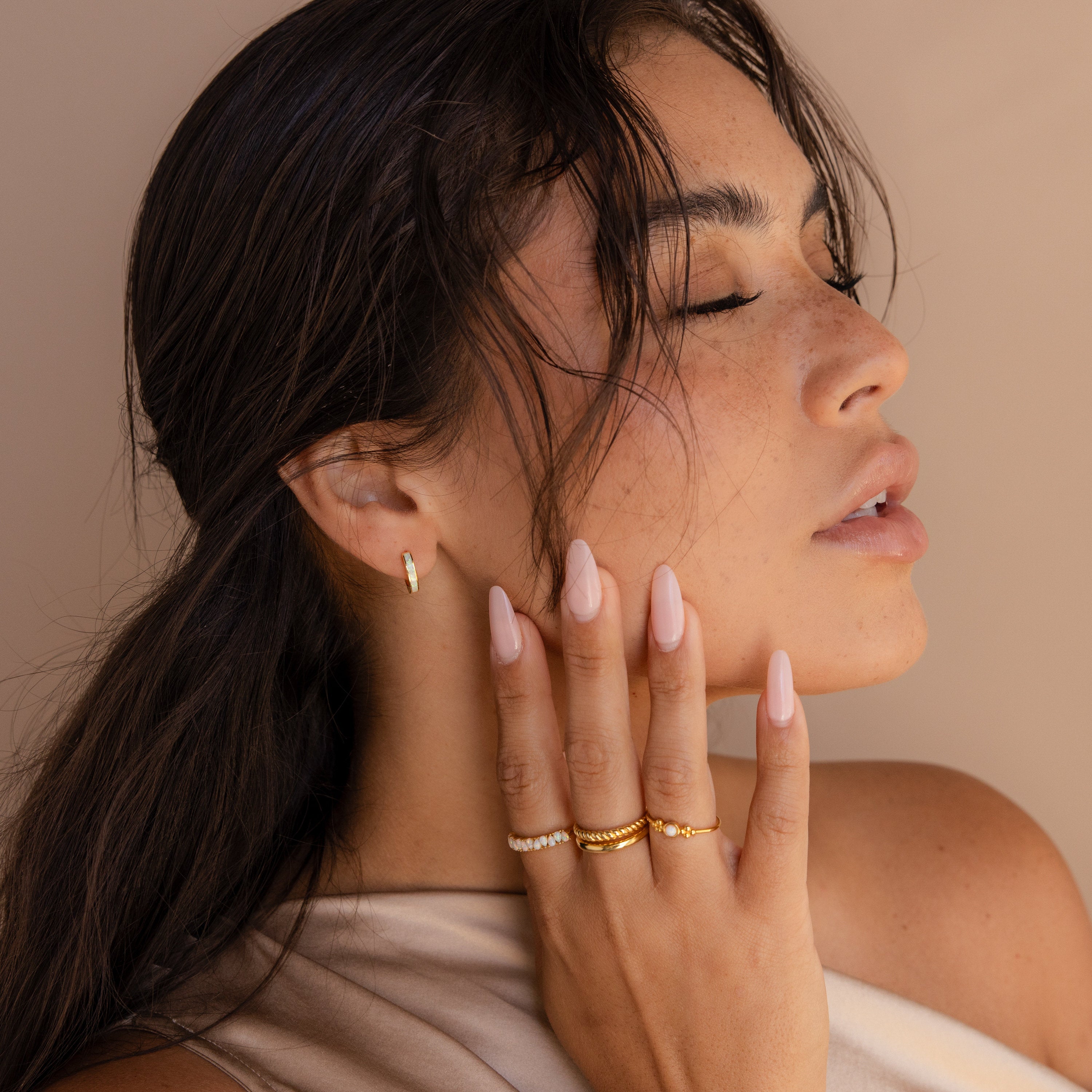 Woman with long nude nails, gold rings, and White Opal Inlay Huggies touches her face with eyes closed against a beige background.