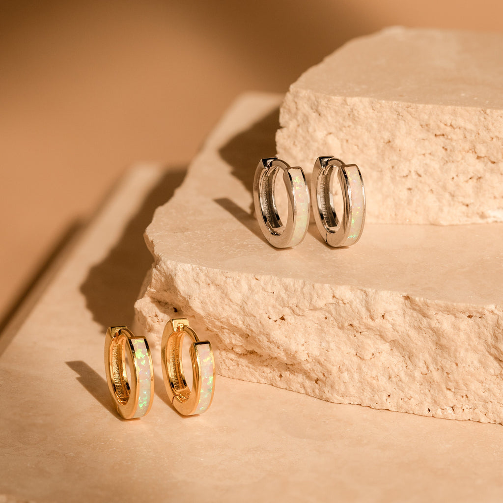 A pair of White Opal Inlay Huggies rests elegantly on a stone surface, showcasing shimmering opal accents that catch the light beautifully.