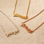 Three Retro Name Necklace in Box Chain pendants—Hannah, Natalie, and Scorpio—are displayed on textured beige fabric, highlighting the timeless appeal of personalized jewelry.