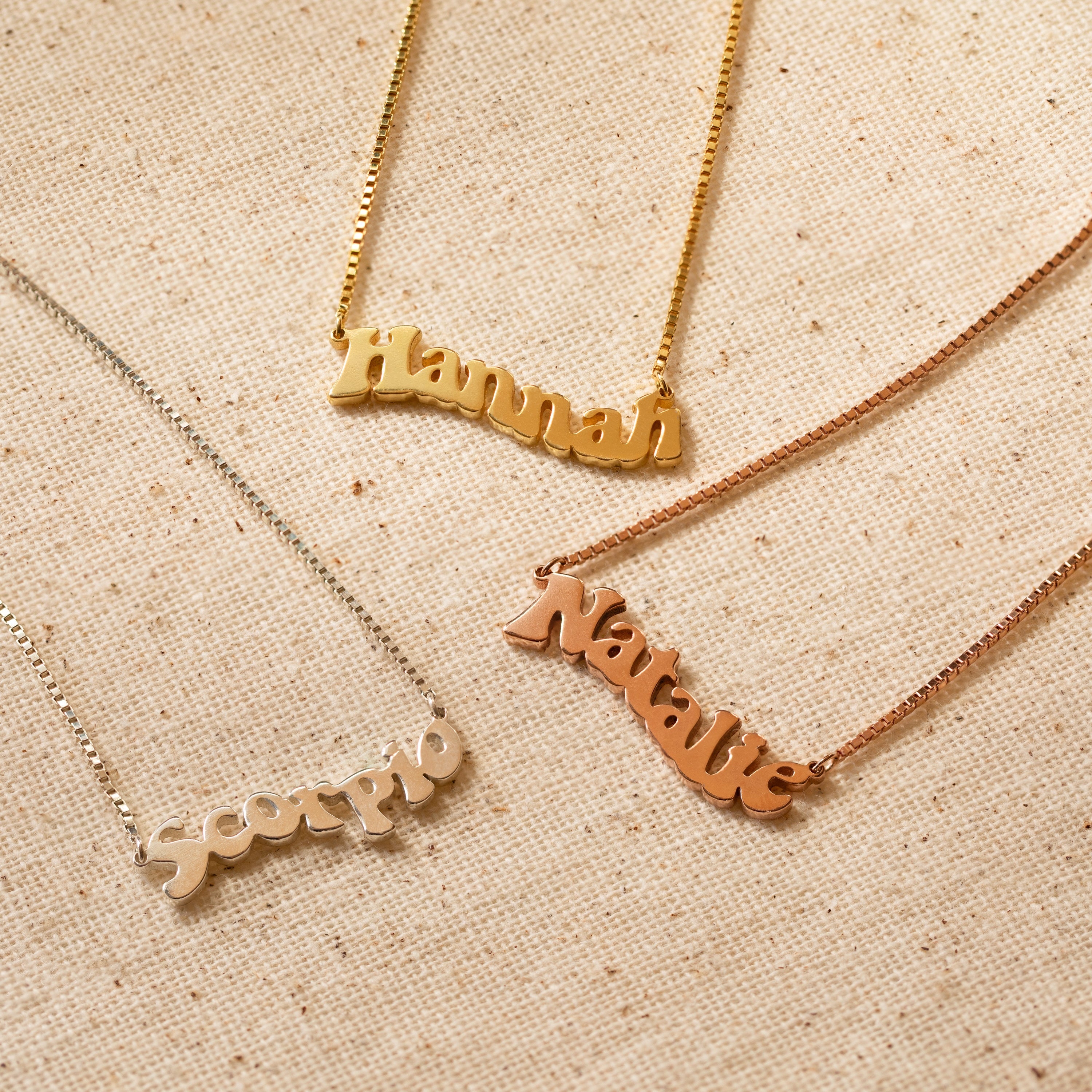 Three Retro Name Necklace in Box Chain pendants—Hannah, Natalie, and Scorpio—are displayed on textured beige fabric, highlighting the timeless appeal of personalized jewelry.