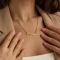A woman wearing the Retro Name Necklace in Box Chain that reads "Amanda," styled with a beige blazer and manicured pink nails.