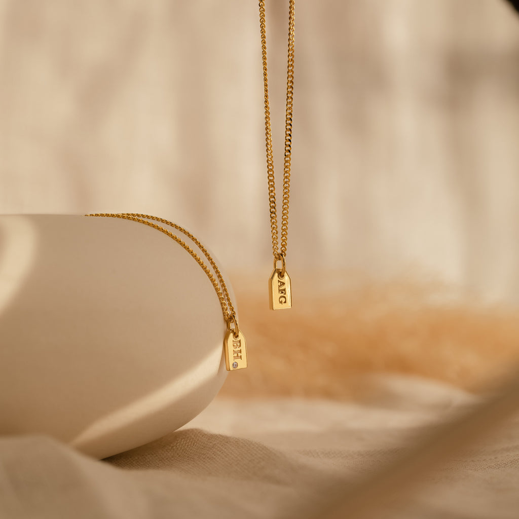 The Tiny Initial Tag Necklace features two gold necklaces with small engraved rectangular pendants, perfect for personalization, shown on a neutral fabric background.