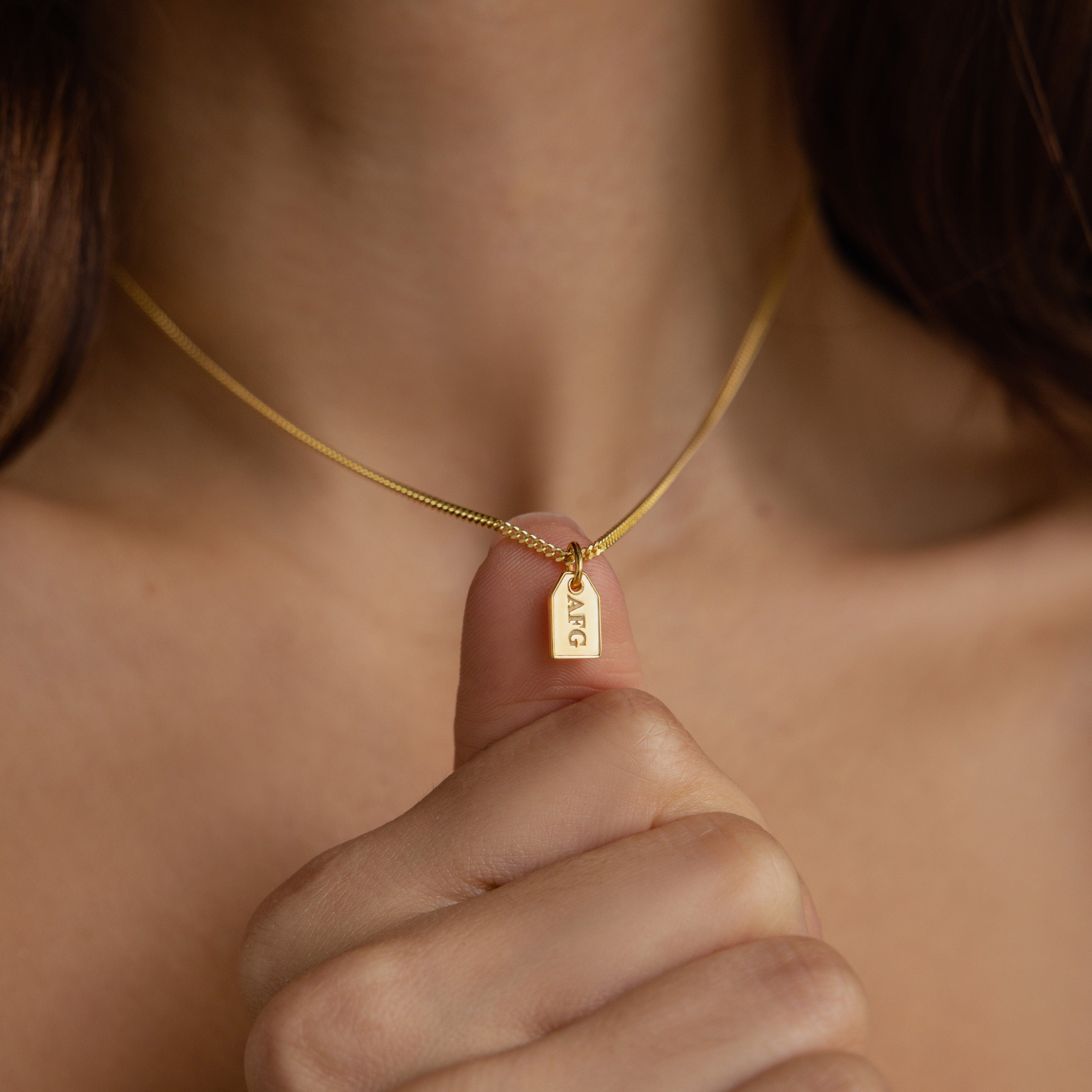 A hand holds the Tiny Initial Tag Necklace, showcasing a gold rectangular pendant engraved with “AGC” for a delicate, personalized touch to any look.