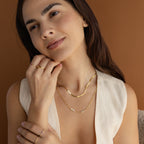 A woman with long brown hair smiles gently, wearing gold necklaces, including the Mini Name Birthstone Necklace, and rings, with her hand near her face.