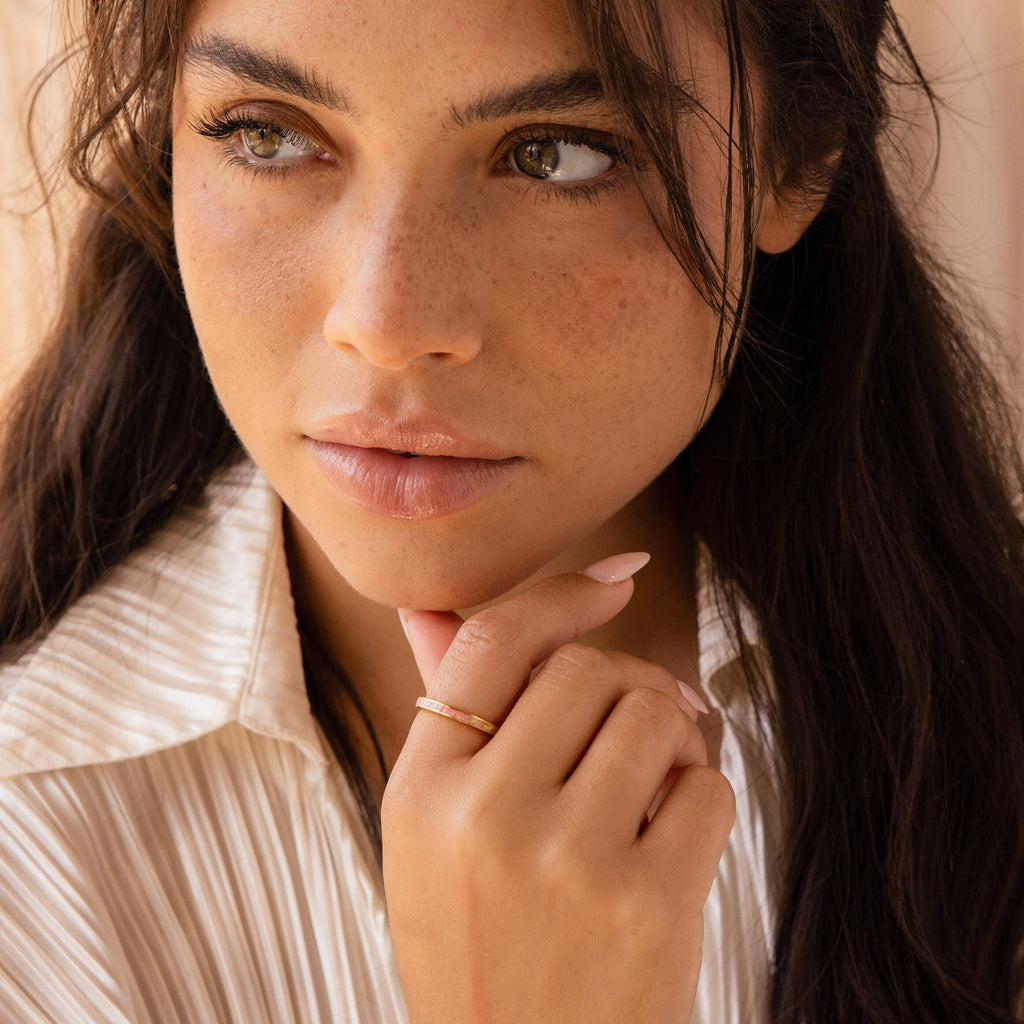 Woman with long dark hair and freckles, wearing a cream blouse and the Pink Opal Inlay Band, looks thoughtfully to the side.