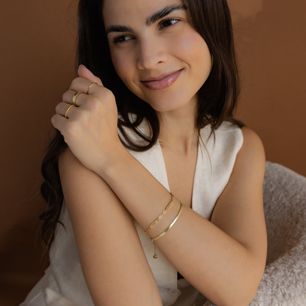 A smiling woman wears the Mini Name Birthstone Bracelet and gold rings, paired with a sleeveless cream top, set against a brown background.