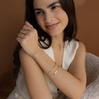 A smiling woman wears the Mini Name Birthstone Bracelet and gold rings, paired with a sleeveless cream top, set against a brown background.