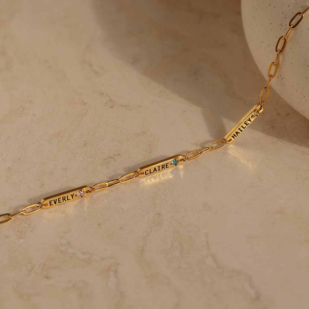 The Mini Name Birthstone Bracelet, featuring engraved name plates reading EVERLY, CLAIRE, and WHALEY, rests on a light marble surface—a perfect customizable gift for someone special.