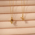 The Initial Heart Birthstone Necklace and two gold necklaces with "Me" pendants are displayed against a soft, peach-colored textured background.