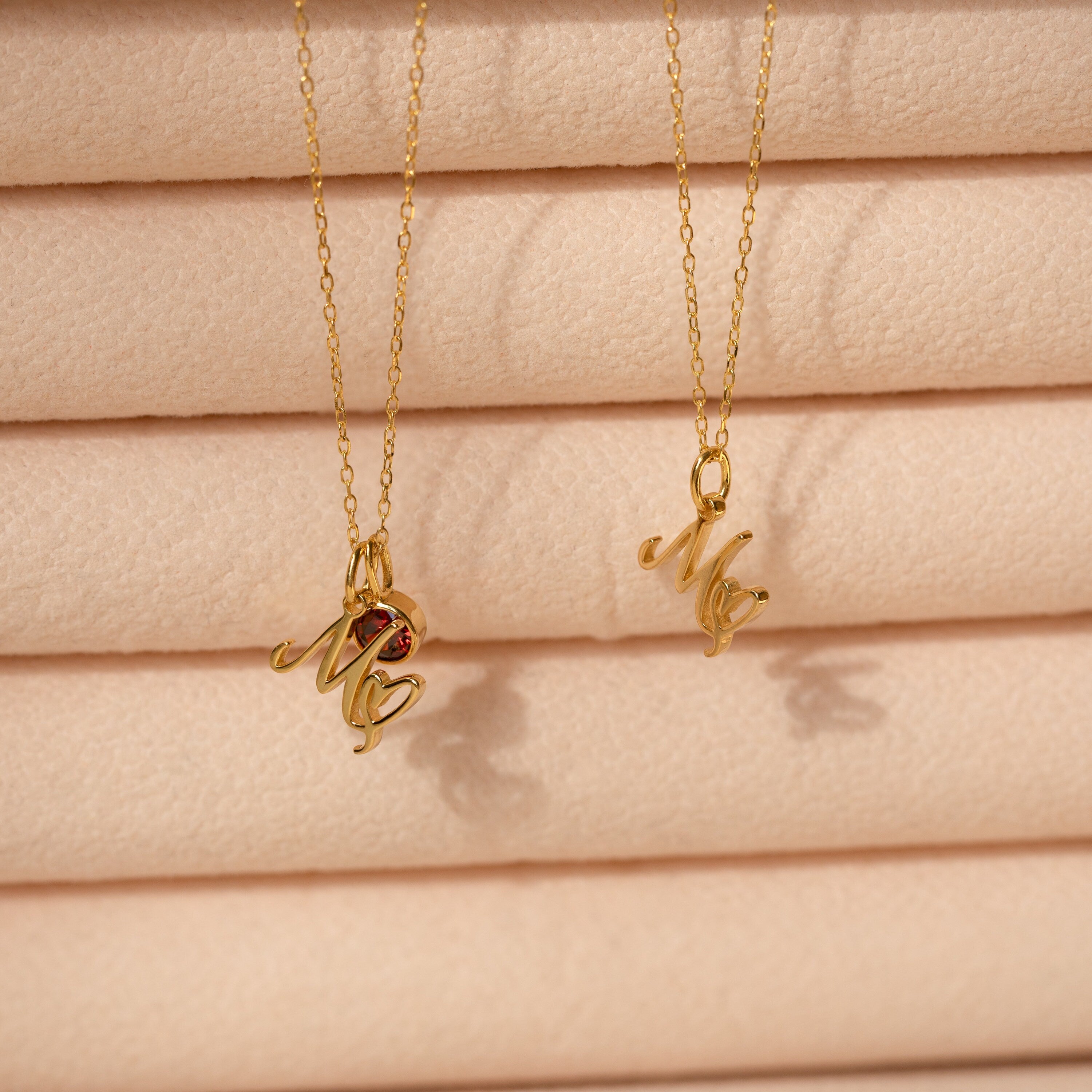 The Initial Heart Birthstone Necklace and two gold necklaces with "Me" pendants are displayed against a soft, peach-colored textured background.
