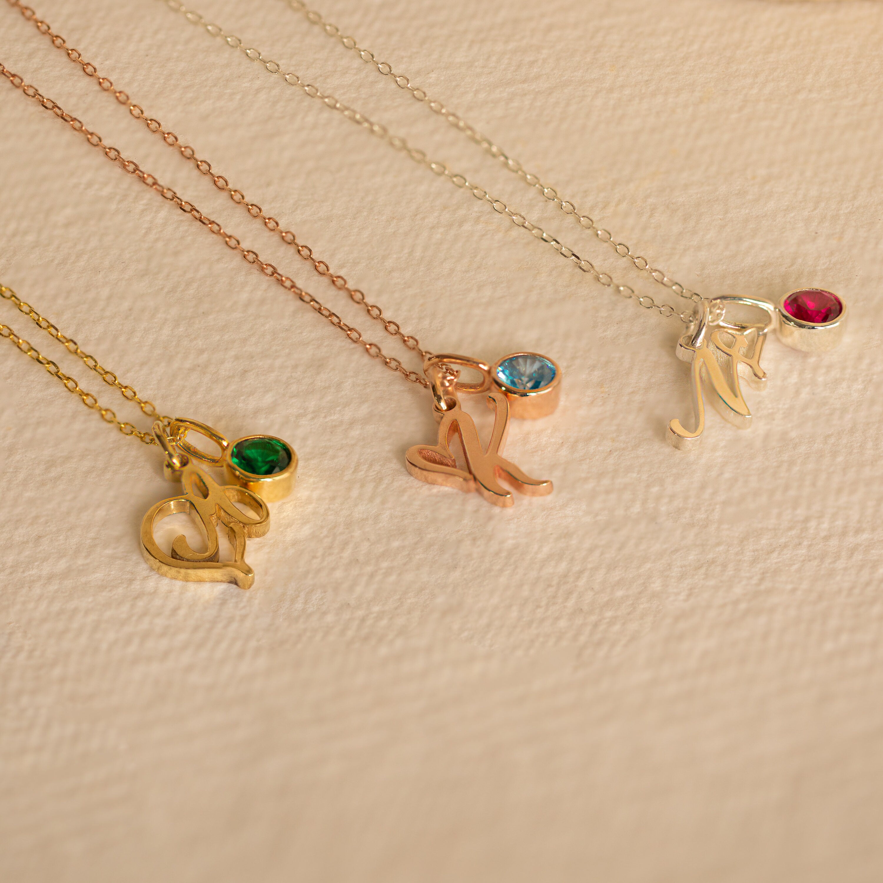 Three Initial Heart Birthstone Necklaces, each with an "M" letter pendant and a custom birthstone charm, are displayed on a textured beige background.
