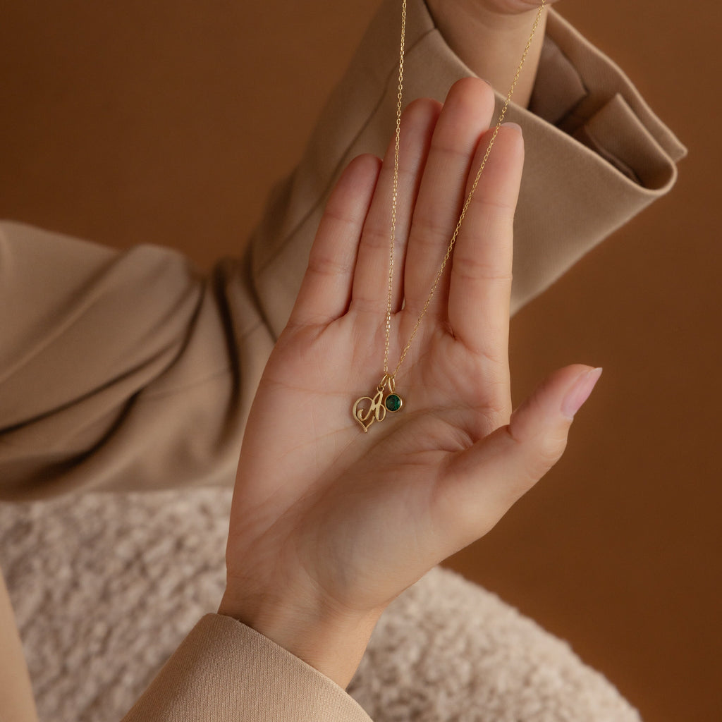 A hand in a beige sleeve holds the Initial Heart Birthstone Necklace, featuring delicate green and gold pendants.