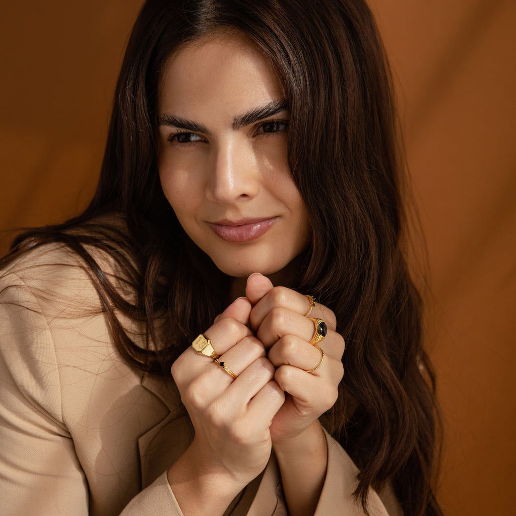A woman with long brown hair, in a beige jacket and bold gold jewelry—including the Lucky Number Signet Ring—smiles softly at the camera.