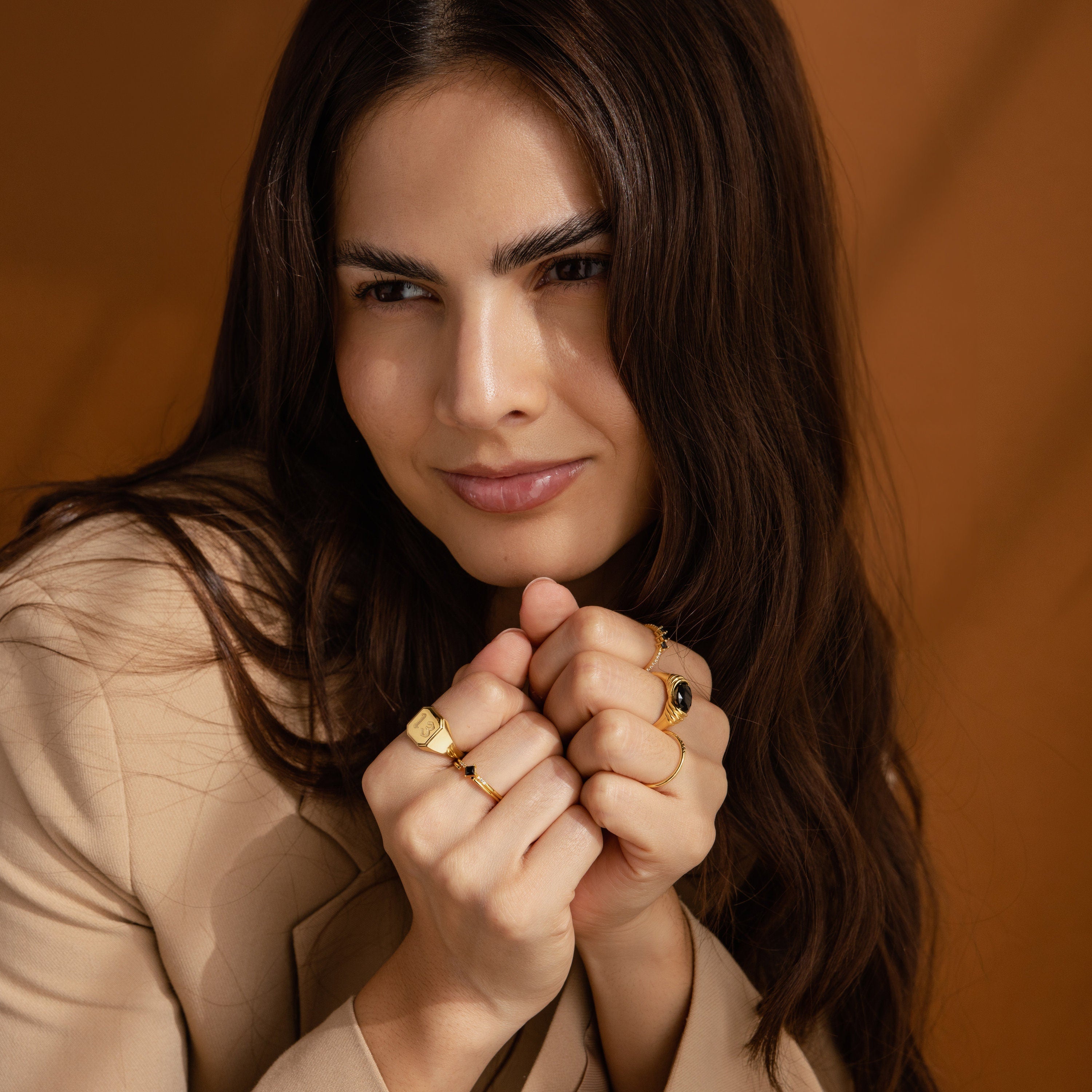 A woman with long brown hair, in a beige jacket and bold gold jewelry—including the Lucky Number Signet Ring—smiles softly at the camera.