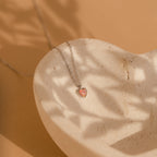 The Pink Opal Heart Necklace, featuring a delicate silver chain, is elegantly displayed in a stone dish on a beige background, highlighting the opal's soft beauty.