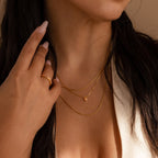 A woman with long dark hair in a cream top touches her neck, showing off dainty gold jewelry and the Pink Opal Heart Necklace for an elegant look.