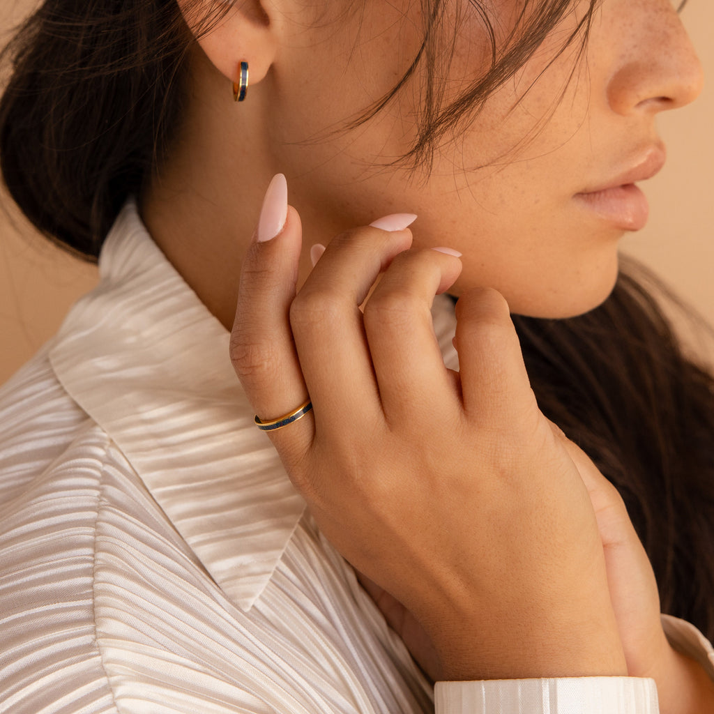 A woman with long nails wears gold jewelry, including the Indigo Opal Inlay Band, a pleated ivory blouse, and gently touches her face.
