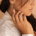 A woman with long nails wears gold jewelry, including the Indigo Opal Inlay Band, a pleated ivory blouse, and gently touches her face.